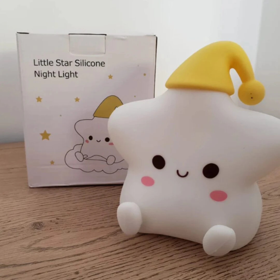 Lámpara de noche con forma de estrella en silicona blanca, tiene cara sonriente, mejillas rosadas y un gorro de dormir amarillo, junto a su caja original.