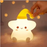 Lámpara nocturna de silicona suave en forma de estrella sonriente con gorro amarillo, luz cálida y difusa, sostenida por una mano en un entorno acogedor.