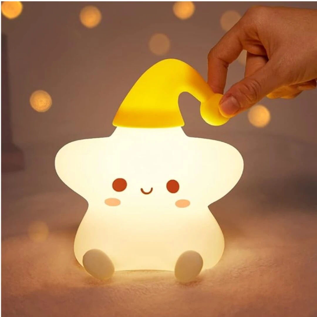 Lámpara nocturna de silicona suave en forma de estrella sonriente con gorro amarillo, luz cálida y difusa, sostenida por una mano en un entorno acogedor.