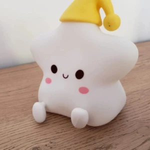 Lámpara nocturna de silicona en forma de estrella blanca sentada, con gorro amarillo, ojos expresivos y mejillas rosadas sobre una superficie de madera.