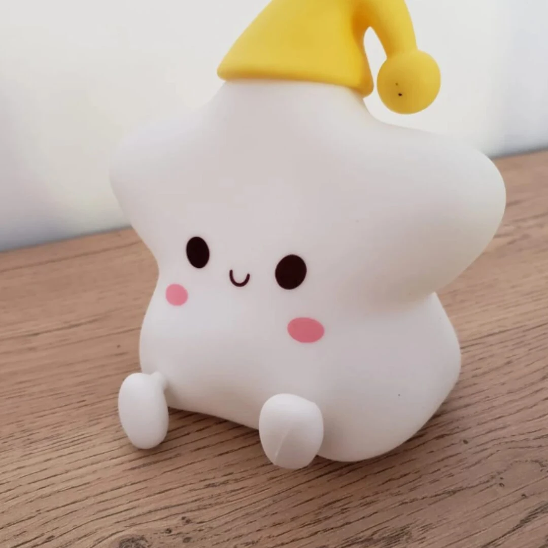 Lámpara nocturna de silicona en forma de estrella blanca sentada, con gorro amarillo, ojos expresivos y mejillas rosadas sobre una superficie de madera.