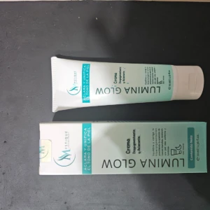 Crema despigmentante Lumina Glow en tubo blanco y su caja protectora, ambos con diseño en tonos verde menta y blanco, contenido neto de 70 ml.