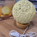 Lámpara de mesa circular con pantalla de hilos entrelazados color crema, base de madera clara, cable USB con interruptor y caja de empaque al fondo.