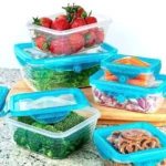 Conjunto de contenedores plásticos transparentes con tapas herméticas azul turquesa, almacenando fresas, brócoli, cebolla, zanahoria y pretzels en cocina.