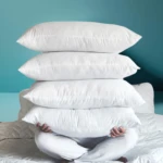 Persona sentada en una cama sosteniendo una torre de cuatro almohadas siliconadas blancas, con textura suave y acabado en color blanco sólido.