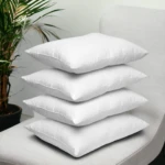 Pila de cuatro almohadas siliconadas de color blanco con acabado suave, dispuestas verticalmente sobre un sillón gris al lado de una planta de interior.