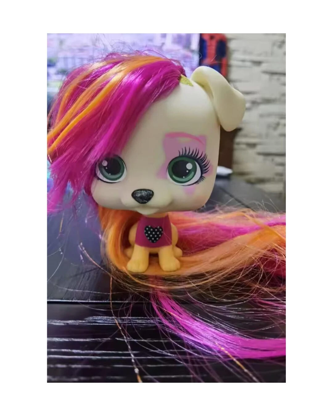 Mascota VIP de juguete con cabeza grande, ojos verdes y cabello largo de colores fucsia y naranja, con un diseño de corazón en su camiseta y maquillaje.
