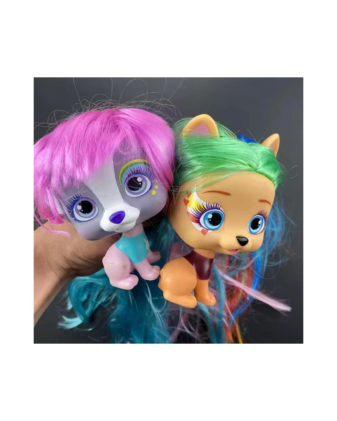 Dos muñecas tipo mascota VIP con cabello largo y colorido, una con pelo rosado y otra con pelo verde, sostenidas por una mano sobre fondo negro.
