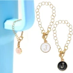 Dos pulseras doradas con dije de inicial, una 'S' rosa y dos 'J', una blanca y otra negra.