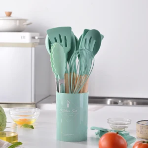 Set de 12 utensilios de cocina en silicona verde menta con mangos de madera, organizados en un recipiente cilíndrico a juego sobre una mesa de cocina.