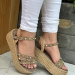 Sandalias de plataforma en yute beige con tiras decoradas con tachuelas doradas, cierre de hebilla y ajuste al tobillo, lucidas con pantalón blanco.