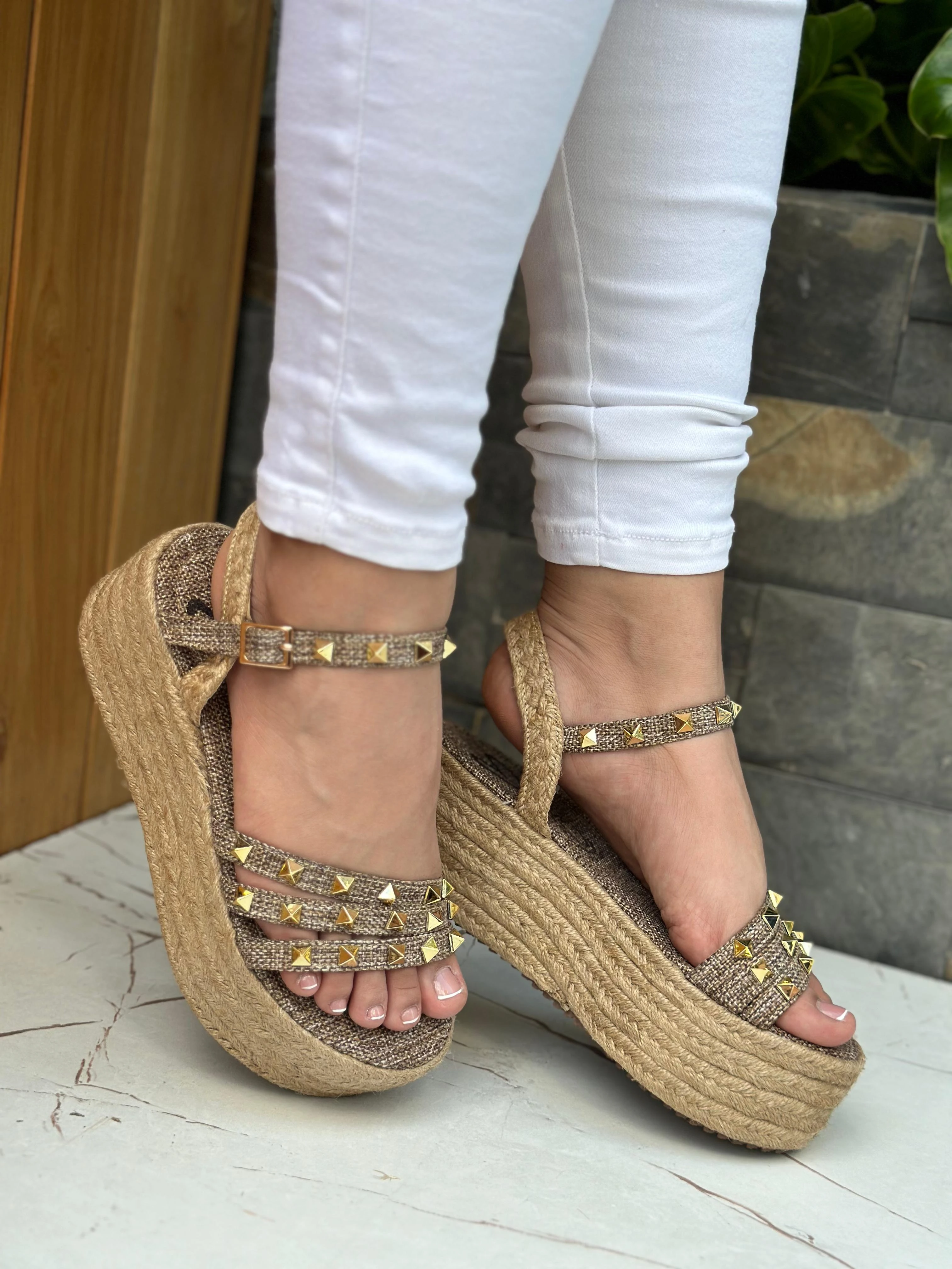 Sandalias de plataforma en yute beige con tiras decoradas con tachuelas doradas, cierre de hebilla y ajuste al tobillo, lucidas con pantalón blanco.