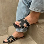 Sandalias planas negras con diseño geométrico y detalles en pedrería brillante, puestas con jeans, sobre una superficie de concreto escalonada.