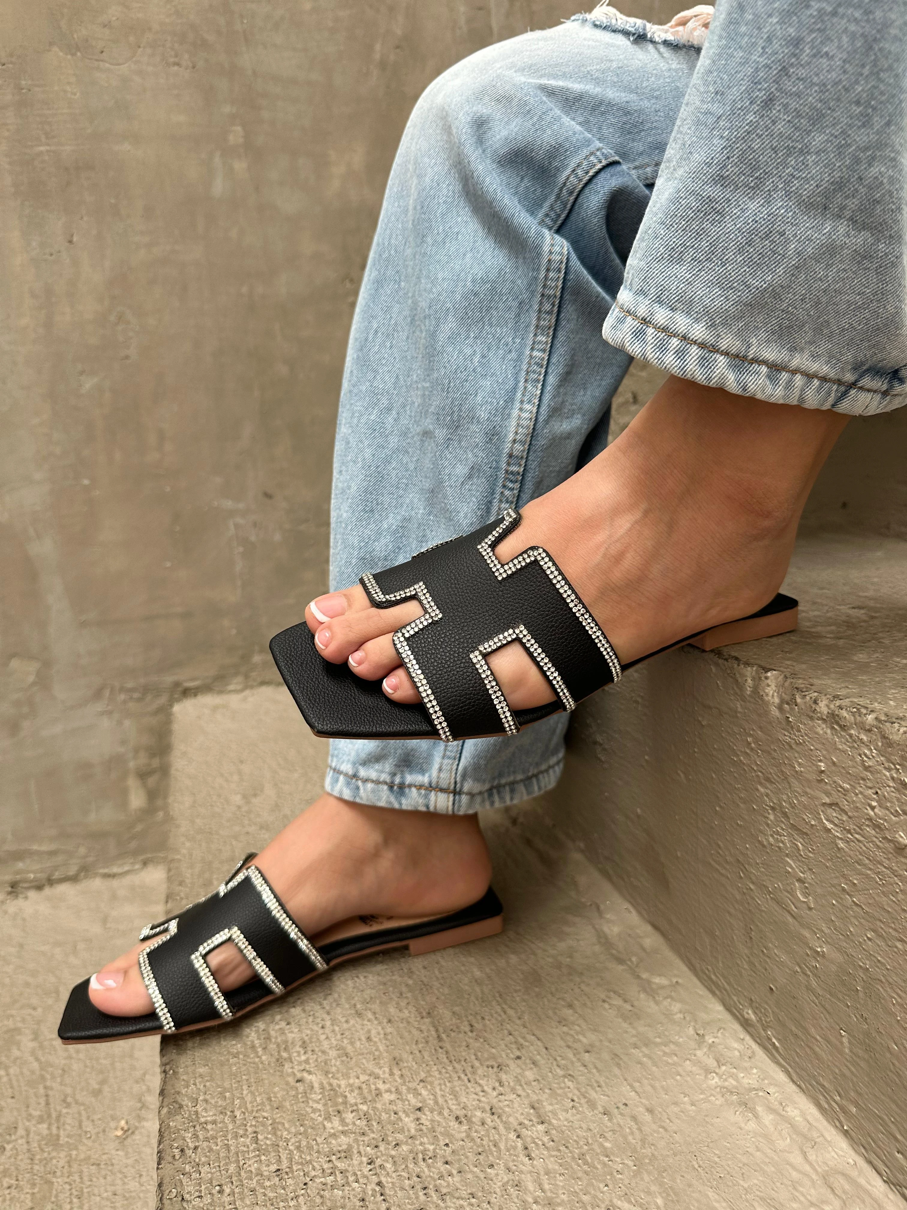 Sandalias planas negras con diseño geométrico y detalles en pedrería brillante, puestas con jeans, sobre una superficie de concreto escalonada.