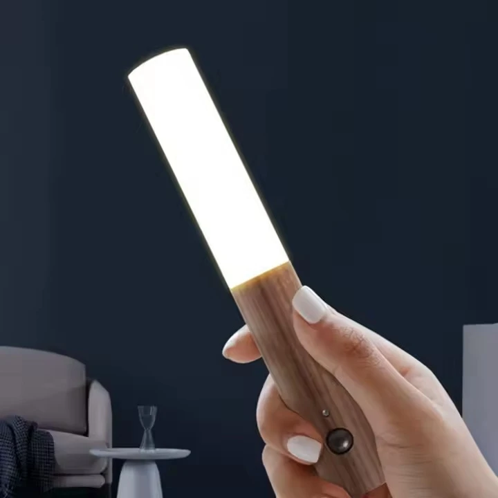 Lámpara portátil LED con cuerpo de acabado madera, sensor de movimiento y luz blanca, sostenida por una mano sobre fondo de sala minimalista.