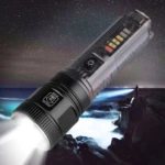Linterna LED negra con indicador de batería y zoom, iluminando paisaje nocturno.