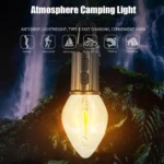 Lámpara de camping tipo bombilla led con gancho de sujeción, cuerpo metálico oscuro, puerto de carga y luz cálida decorativa sobre fondo de cascada.