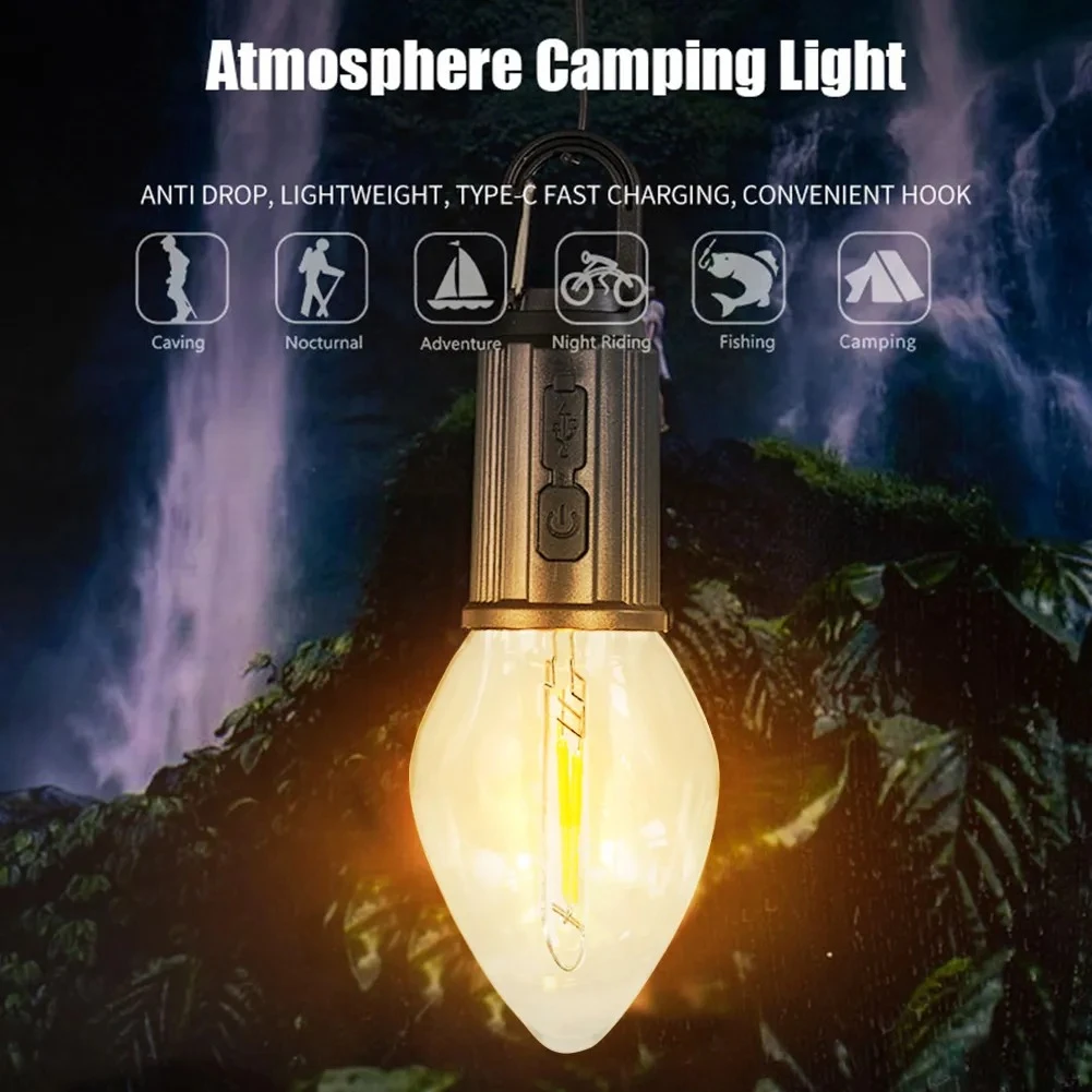 Lámpara de camping tipo bombilla led con gancho de sujeción, cuerpo metálico oscuro, puerto de carga y luz cálida decorativa sobre fondo de cascada.