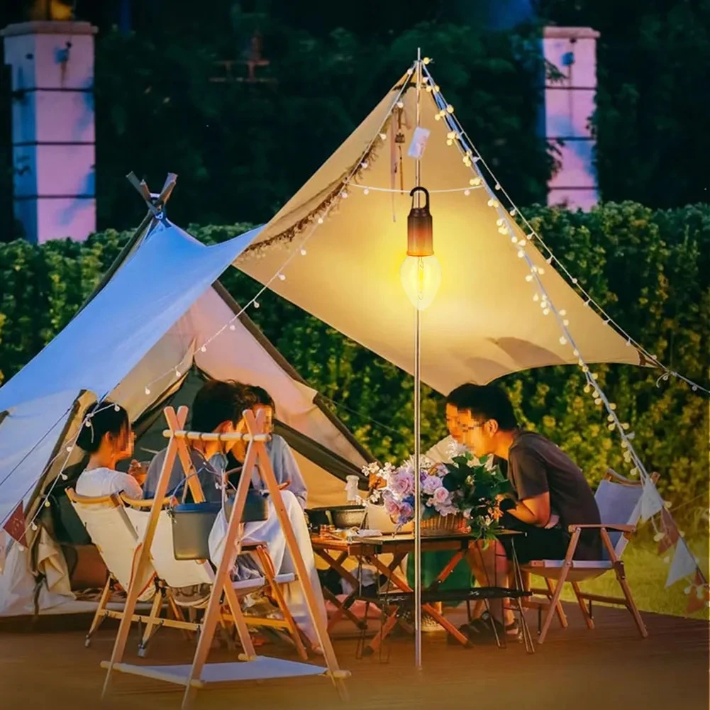 Lámpara LED de camping estilo vintage con base de metal, iluminando una mesa de exterior bajo una carpa tipo toldo con personas reunidas en la noche.