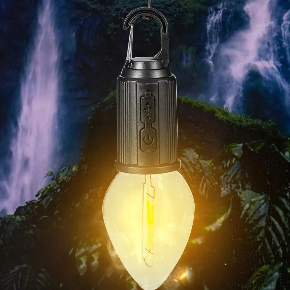 Lámpara de camping LED con forma de bombilla vintage, cuerpo negro mate, gancho tipo mosquetón, puerto USB y botón de encendido, sobre fondo de cascada.