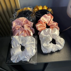 Set de tres scrunchies de satín en colores dorado, negro, rosa y blanco, organizados en parejas sobre una superficie negra, ideales para estilizar el cabello.