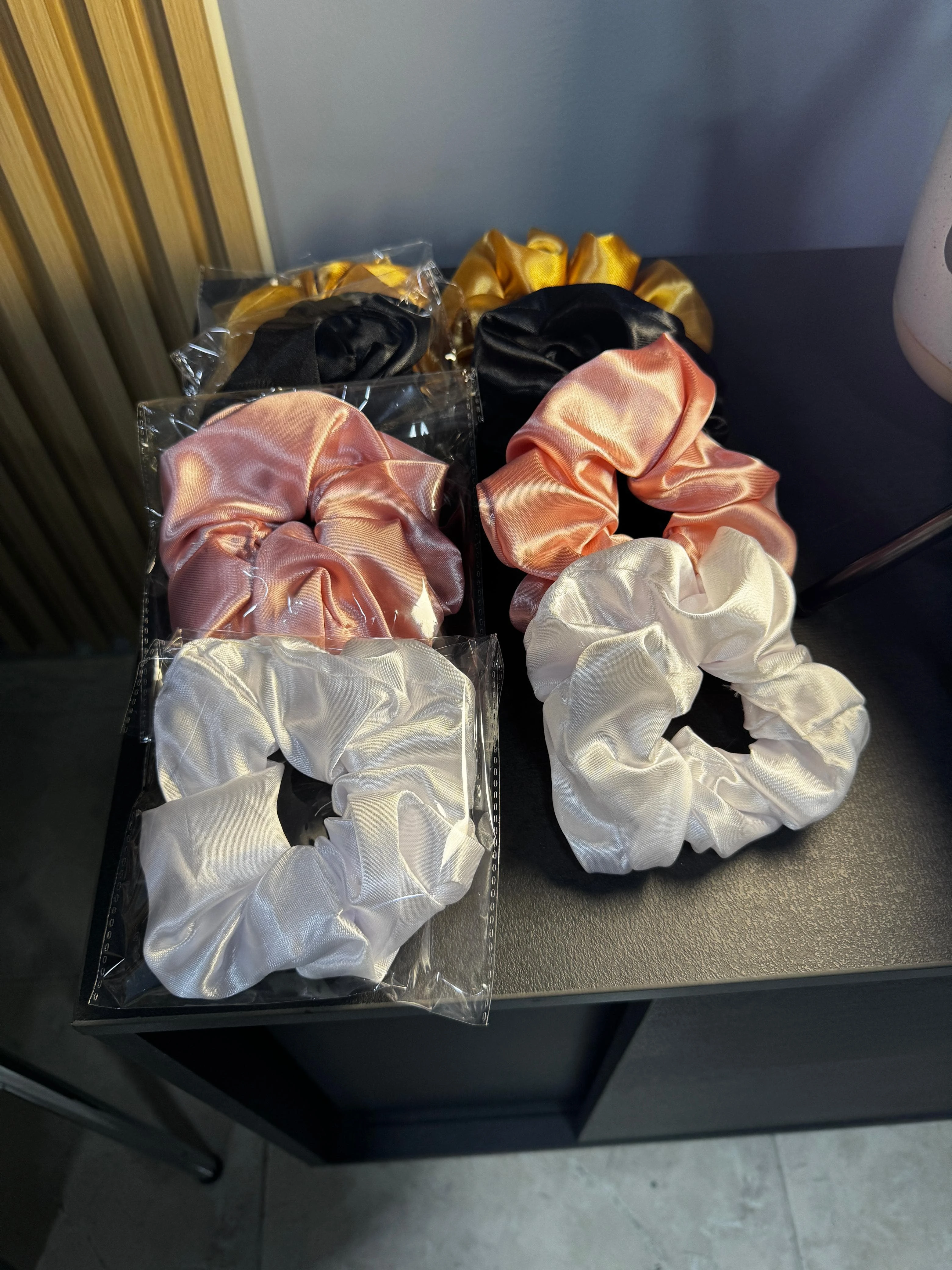 Set de tres scrunchies de satín en colores dorado, negro, rosa y blanco, organizados en parejas sobre una superficie negra, ideales para estilizar el cabello.