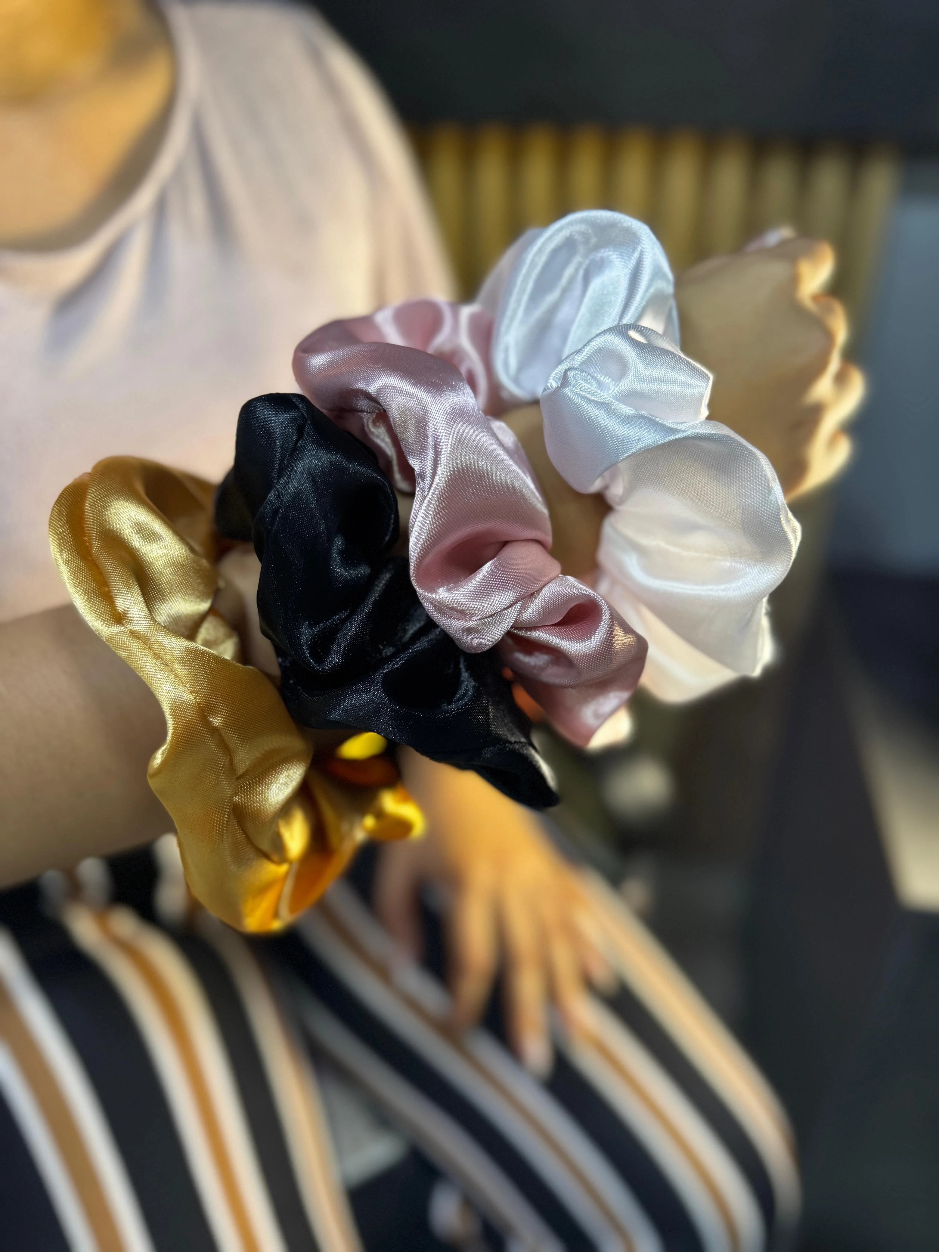Muñeca de mujer con cuatro scrunchies de satén en colores blanco, rosa, negro y dorado, exhibidos para resaltar su textura brillante y acabado elegante.
