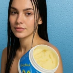 Mujer con cabello mojado y mascarilla capilar Hydromask en tarro blanco y azul, fondo azul con gotas.