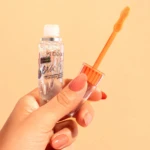 Mano sosteniendo un envase de serum para pestañas Kj Exactitud transparente con aplicador de cerdas naranja, sobre fondo color crema.