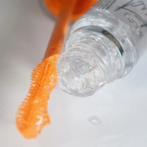 Serum para crecimiento de pestañas con aplicador de cerdas flexibles color naranja, mostrado fuera de su envase transparente y alargado.