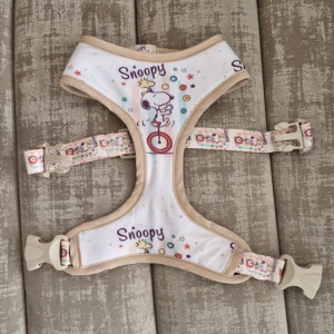 Arnés para perros con estampado de Snoopy, diseño de dos piezas conectadas, correas ajustables con hebillas plásticas color arena y superficie blanca.