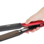 Mano sosteniendo una plancha para cabello Remington de color rojo metalizado con placas negras, abierta y lista para usar, sobre fondo blanco.