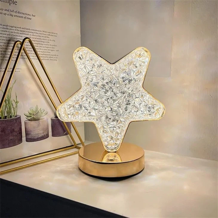 Lámpara de mesa en forma de estrella con superficie facetada transparente y base circular de metal dorado sobre superficie blanca con plantas decorativas.