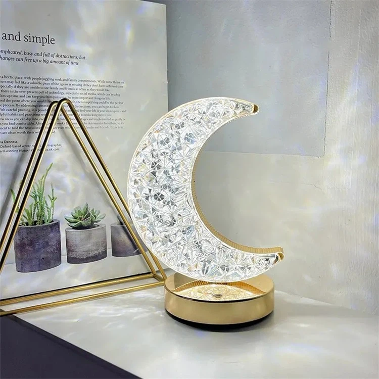 Lámpara de mesa en forma de media luna con textura de cristal tallado, base dorada circular y luz cálida sobre una superficie blanca junto a decoración.