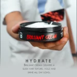 Mano sosteniendo tarro negro de Brilliant Cream Lv3 con crema blanca abierta, resaltando su función hidratante y acabado brillante para peinado.