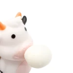 Juguete de silicona blanco con forma de vaca masticando una pelota, detalles naranjas y negros en las orejas.