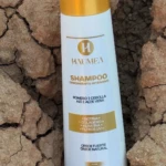 Shampoo blanco Haumea para crecimiento intensivo con tapa dorada sobre fondo de tierra seca agrietada.