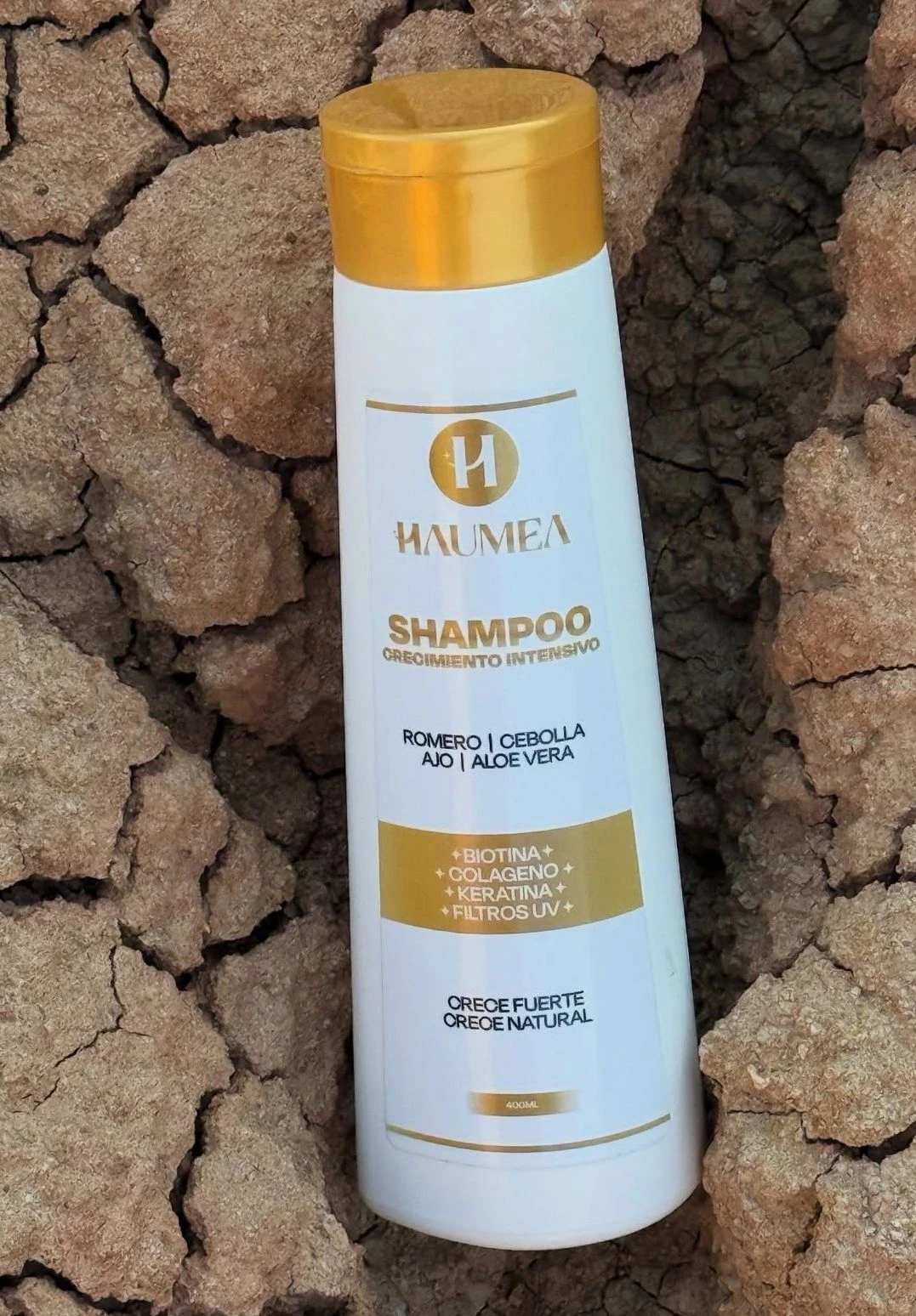 Shampoo blanco Haumea para crecimiento intensivo con tapa dorada sobre fondo de tierra seca agrietada.