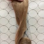 Extensiones de cabello rubio de 60 cm, lisas con puntas onduladas, sujetas por una banda con ganchos y envueltas parcialmente en una red protectora negra.