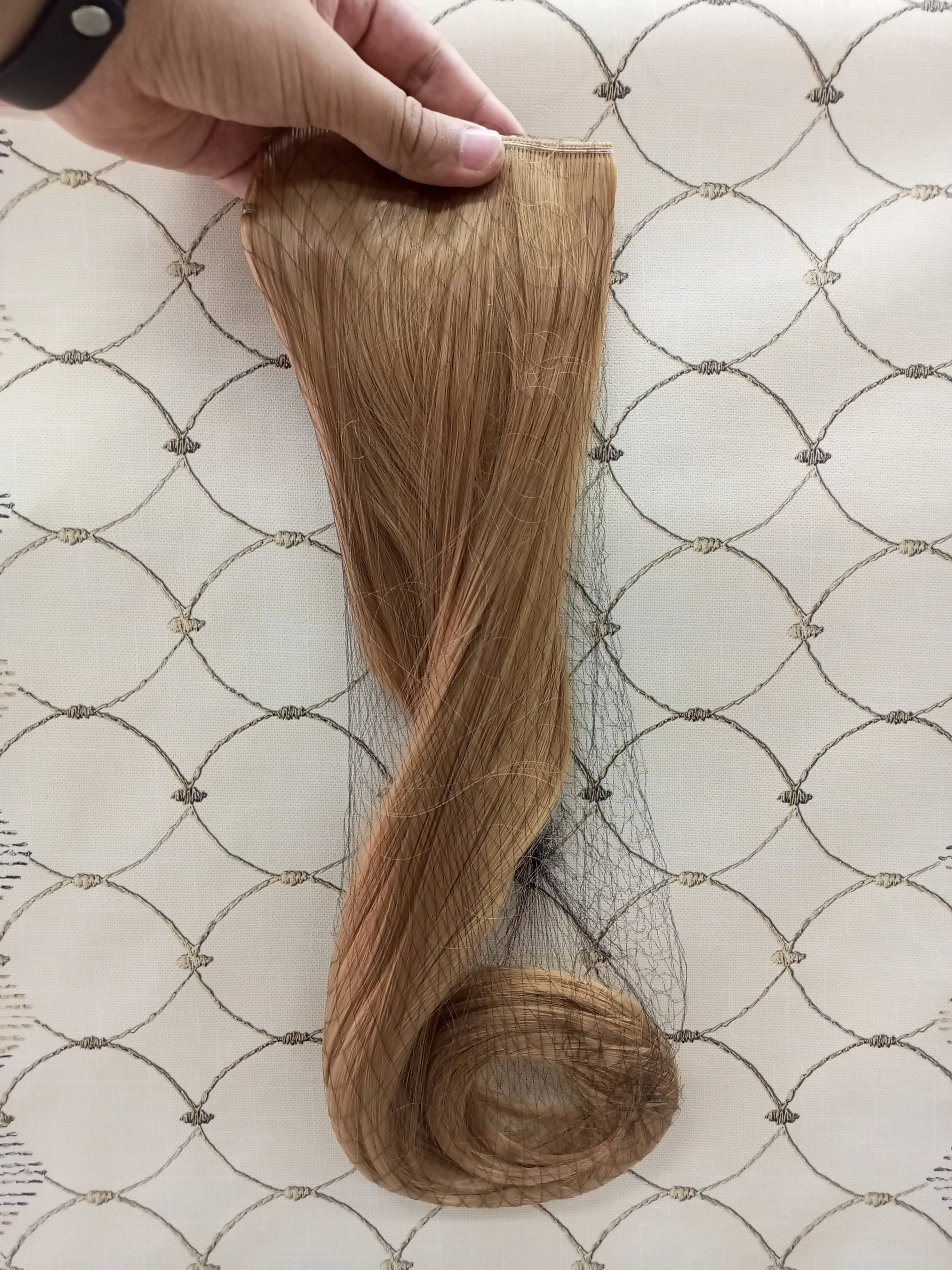 Extensiones de cabello rubio de 60 cm, lisas con puntas onduladas, sujetas por una banda con ganchos y envueltas parcialmente en una red protectora negra.