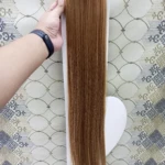 Extensiones de cabello liso color castaño claro, de 60 cm de largo, sujetas a una base con clips, sostenidas por una mano sobre fondo de tela estampada.