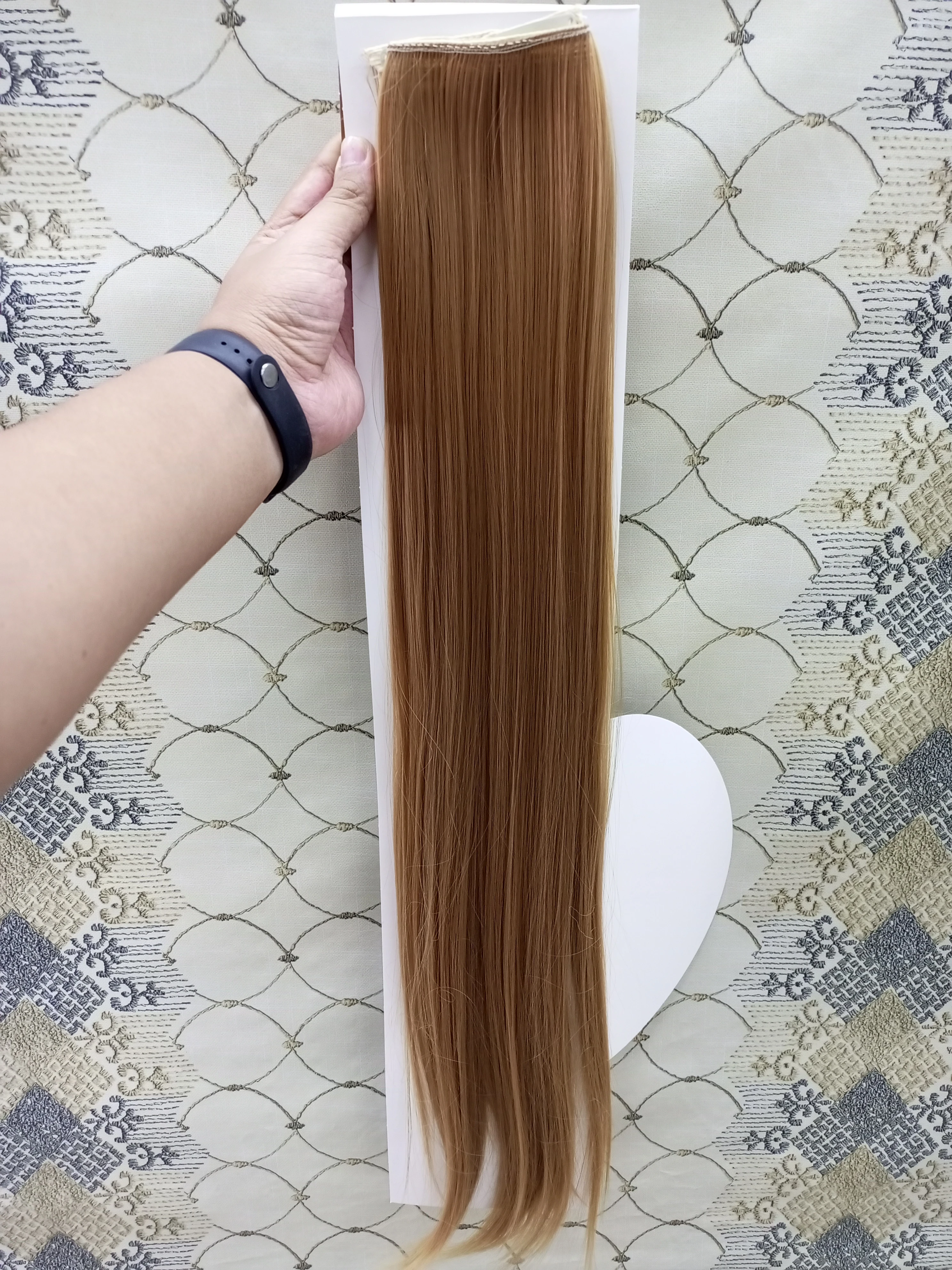 Extensiones de cabello liso color castaño claro, de 60 cm de largo, sujetas a una base con clips, sostenidas por una mano sobre fondo de tela estampada.