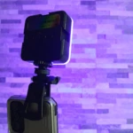 Luz LED RGB para celular, montada en un tripié acoplado a smartphone, con fondo de pared lila.