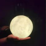 Lámpara LED con forma de luna llena de 15 cm, textura realista de cráteres con luz cálida sostenida sobre una mano en un entorno oscuro.