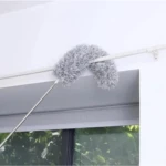 Plumero extensible de microfibra gris curvado limpiando el borde superior de una ventana con marco blanco.