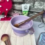 Tazón morado 3 en 1 para limpiar brochas de maquillaje con cepillo de silicona integrado y brocha rosada.