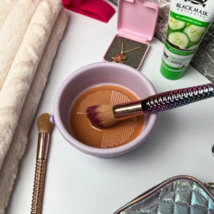 Brocha de maquillaje con mango holográfico en recipiente morado con limpiador y rejilla, toalla suave y crema facial.
