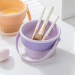 Limpiador de brochas morado y naranja con asas, tres brochas y esponja de maquillaje.