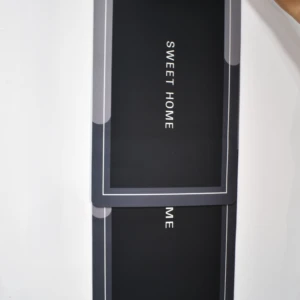 Tapete absorbente negro con diseño "SWEET HOME" y borde gris doble, colgado en pared blanca.