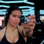 Mujer con auriculares en gimnasio muestra soporte magnético plateado para celular con trípode STTRIKER.
